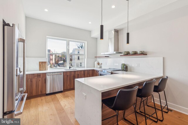 kitchen with brown cabinetry, appliances with stainless steel finishes, a sink, a peninsula, and wall chimney exhaust hood