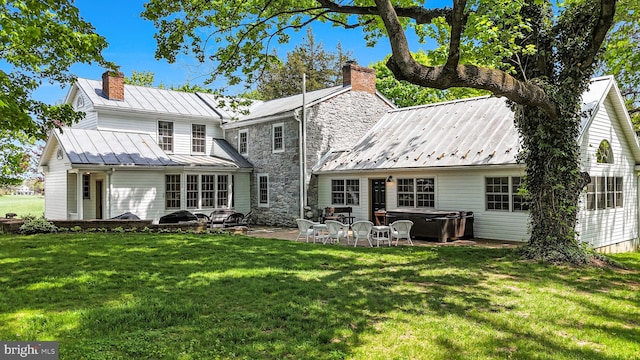 back of house featuring a patio, a chimney, a standing seam roof, a lawn, and a hot tub
