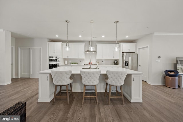 kitchen with wall chimney range hood, appliances with stainless steel finishes, white cabinetry, wood finished floors, and a spacious island