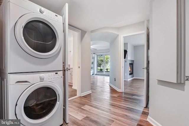 laundry room featuring laundry area, stacked washer and dryer, a glass covered fireplace, baseboards, and light wood-type flooring