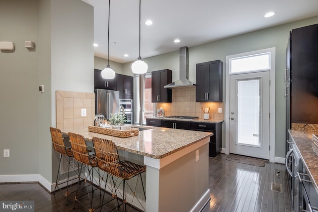 kitchen featuring wall chimney range hood, dark wood-style flooring, light stone countertops, stainless steel appliances, and baseboards