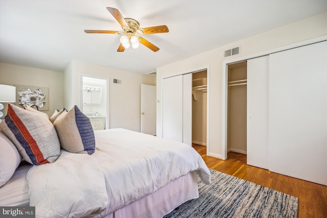 bedroom with two closets, wood finished floors, ensuite bath, and a ceiling fan