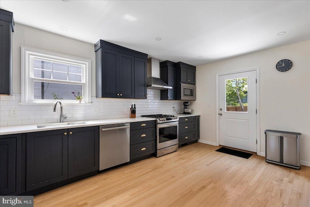 kitchen with a sink, appliances with stainless steel finishes, wall chimney range hood, and light wood finished floors