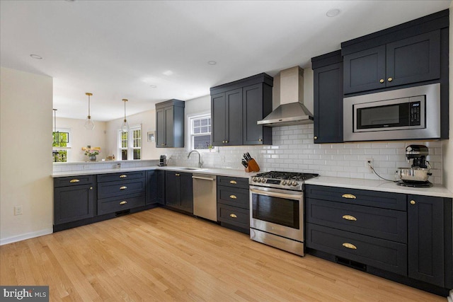 kitchen with wall chimney range hood, backsplash, appliances with stainless steel finishes, light wood-type flooring, and light countertops