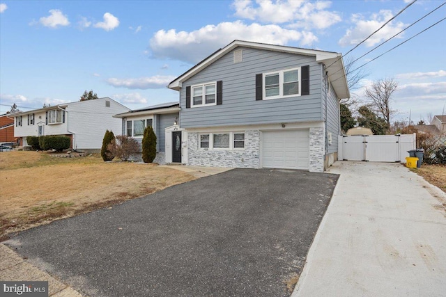 tri-level home featuring a garage, aphalt driveway, fence, stone siding, and a gate