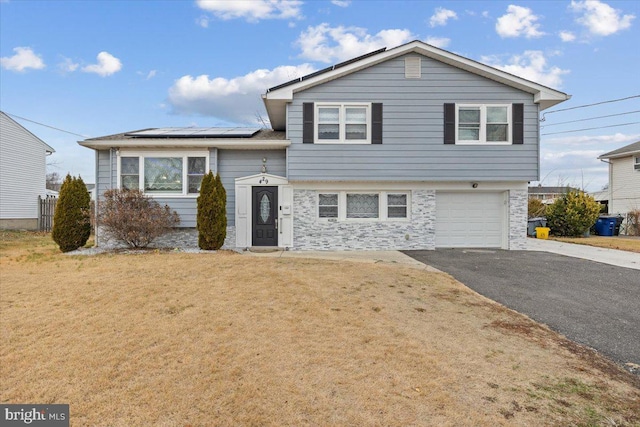 split level home featuring driveway, a front yard, a garage, roof mounted solar panels, and stone siding