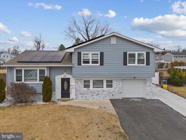 tri-level home featuring driveway, solar panels, a front yard, a garage, and stone siding