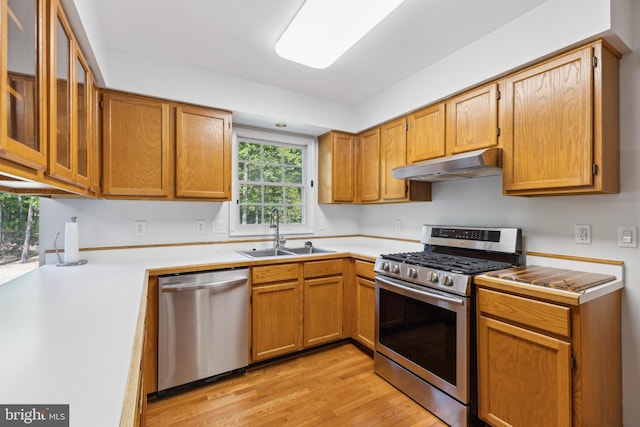 kitchen with appliances with stainless steel finishes, light wood-type flooring, a sink, light countertops, and under cabinet range hood