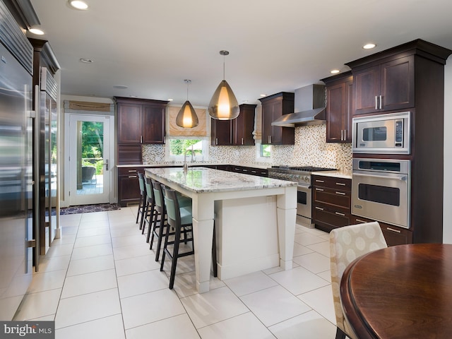 kitchen featuring appliances with stainless steel finishes, a kitchen island with sink, wall chimney exhaust hood, tasteful backsplash, and a kitchen bar