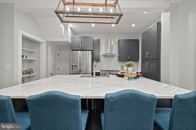kitchen featuring stainless steel refrigerator with ice dispenser, wall chimney range hood, a sink, gray cabinetry, and recessed lighting