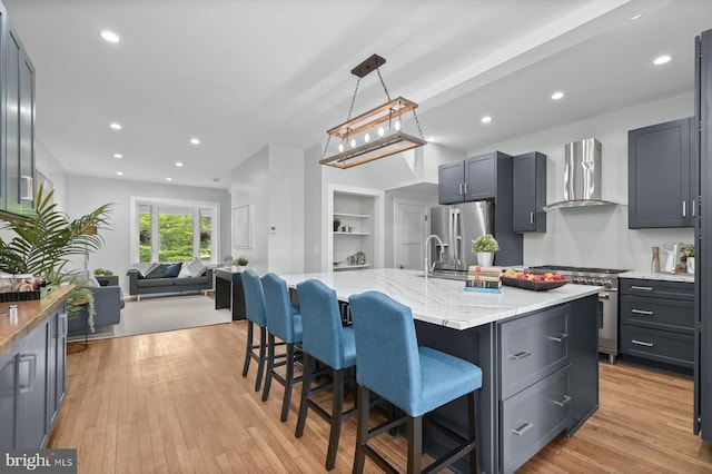 kitchen featuring wall chimney range hood, stainless steel appliances, open floor plan, a kitchen bar, and light wood finished floors