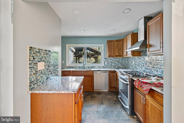 kitchen featuring brown cabinets, appliances with stainless steel finishes, a sink, stone finish floor, and wall chimney exhaust hood