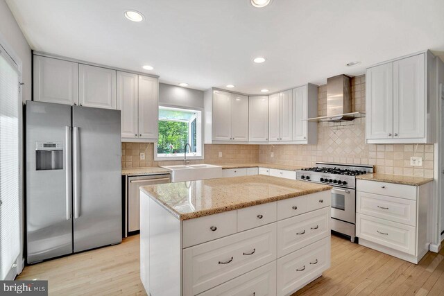 kitchen featuring appliances with stainless steel finishes, wall chimney exhaust hood, light stone countertops, a sink, and light wood-style flooring