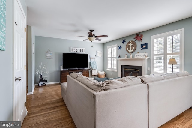 living area featuring a ceiling fan, baseboards, a glass covered fireplace, and wood finished floors