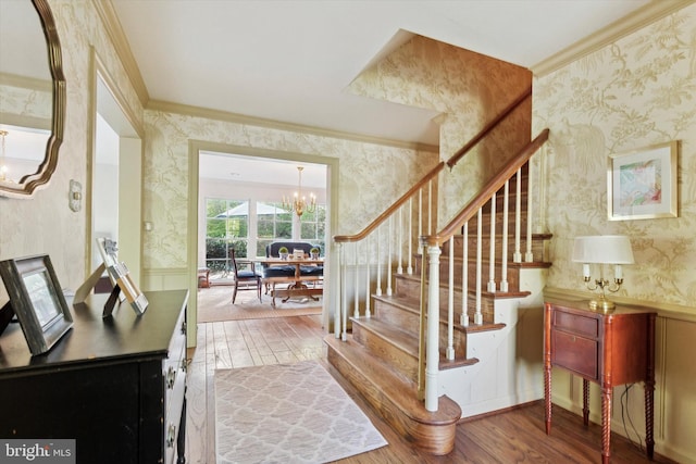 stairway with an inviting chandelier, ornamental molding, wood finished floors, and wallpapered walls