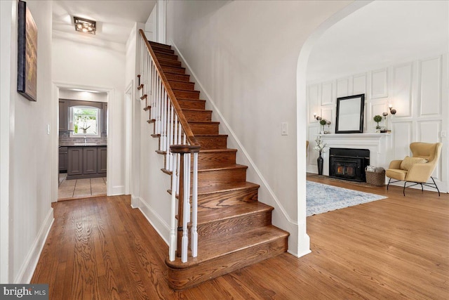 staircase with arched walkways, baseboards, a lit fireplace, and wood finished floors