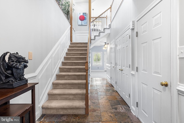 staircase featuring stone tile floors, a decorative wall, a towering ceiling, baseboards, and wainscoting
