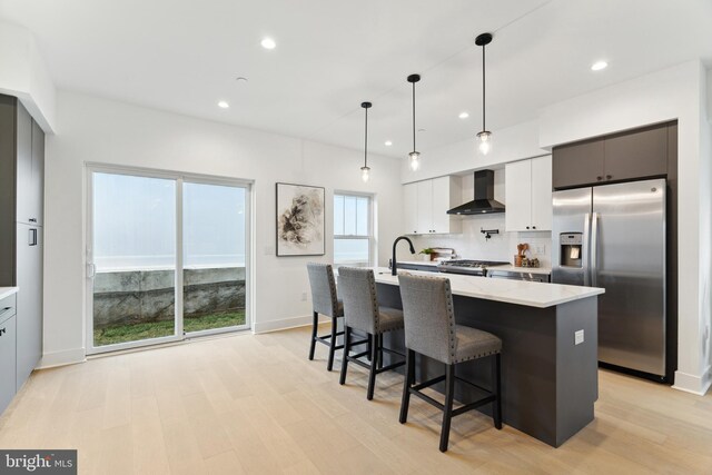 kitchen featuring wall chimney range hood, a kitchen island with sink, backsplash, light wood finished floors, and stainless steel appliances