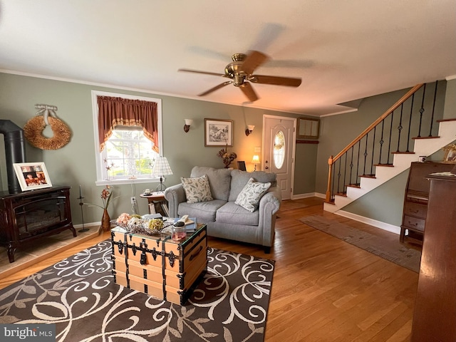living area featuring crown molding, a wood stove, stairs, wood finished floors, and ceiling fan