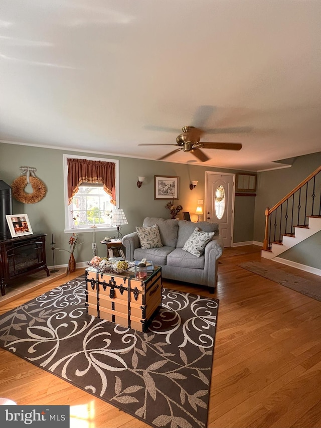 living area with a ceiling fan, a wood stove, stairs, and wood finished floors