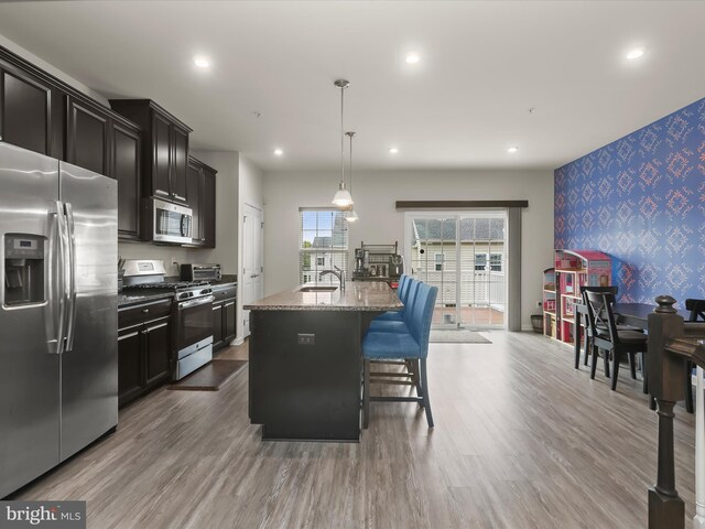 kitchen with stainless steel appliances, wallpapered walls, stone countertops, an accent wall, and an island with sink