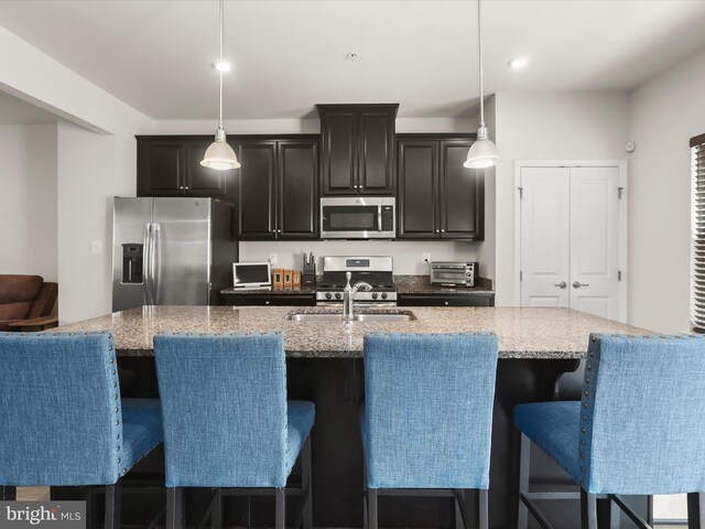 kitchen featuring appliances with stainless steel finishes, a sink, a kitchen island with sink, dark cabinets, and pendant lighting