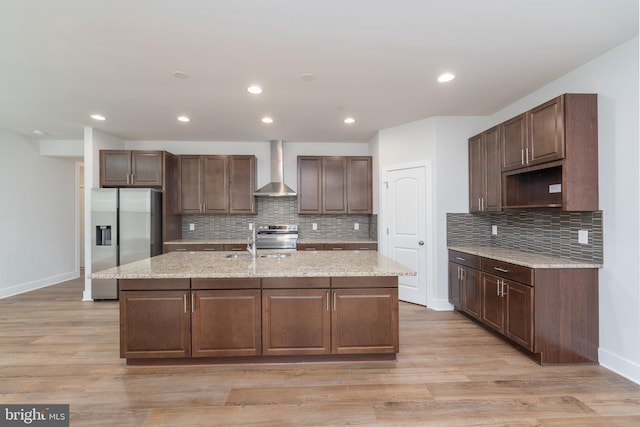 kitchen with wall chimney range hood, a kitchen island with sink, backsplash, baseboards, and stainless steel appliances