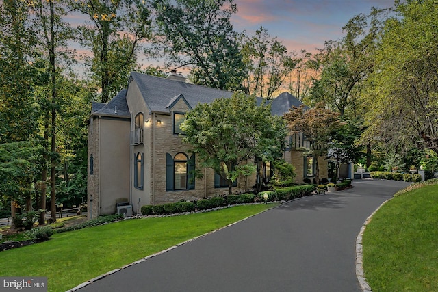 french country style house with a balcony, a front lawn, a shingled roof, stone siding, and stucco siding