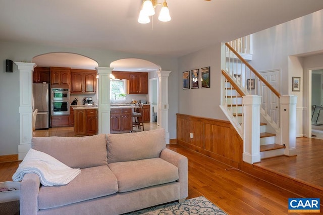 living area featuring wainscoting, light wood-style floors, arched walkways, stairs, and ornate columns