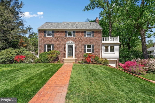 colonial inspired home with a chimney, a garage, brick siding, a balcony, and a front lawn