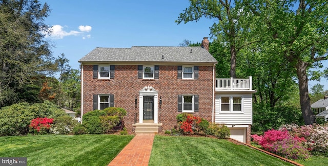 colonial-style house with a chimney, a garage, brick siding, a balcony, and a front lawn