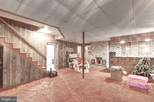 finished basement featuring stairs, a brick fireplace, wooden walls, a paneled ceiling, and carpet flooring