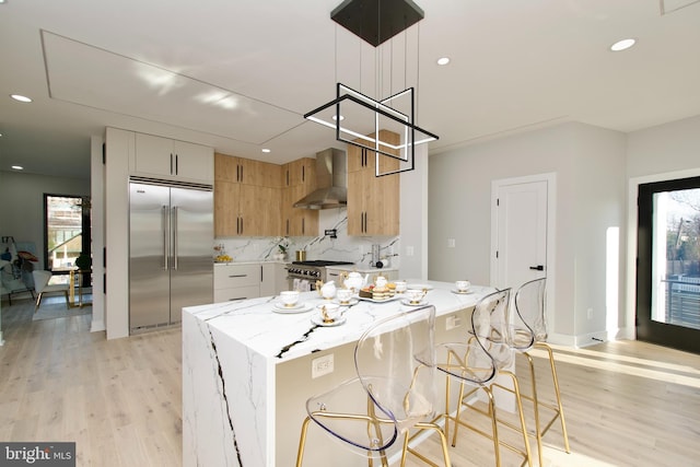 kitchen with decorative backsplash, high end appliances, wall chimney range hood, light stone counters, and light wood-style flooring