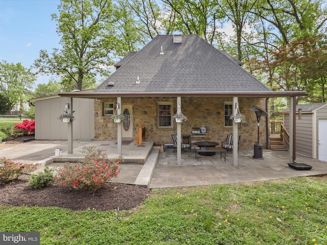 rear view of property featuring stone siding, a patio area, roof with shingles, a shed, and an outbuilding