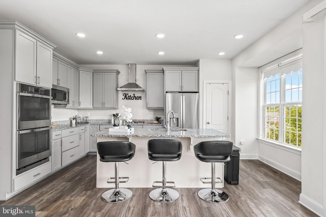 kitchen featuring light stone countertops, gray cabinetry, a sink, stainless steel appliances, and wall chimney exhaust hood