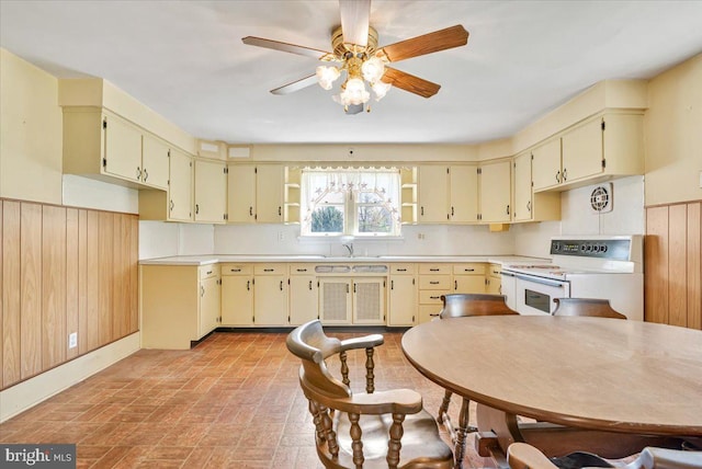 kitchen with ceiling fan, wood walls, a wainscoted wall, cream cabinetry, and white electric range