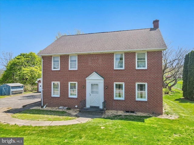 colonial-style house with driveway, a chimney, a front yard, brick siding, and a shingled roof