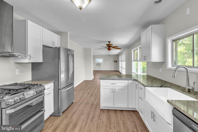 kitchen featuring wall chimney range hood, appliances with stainless steel finishes, ceiling fan, light wood-style floors, and a sink