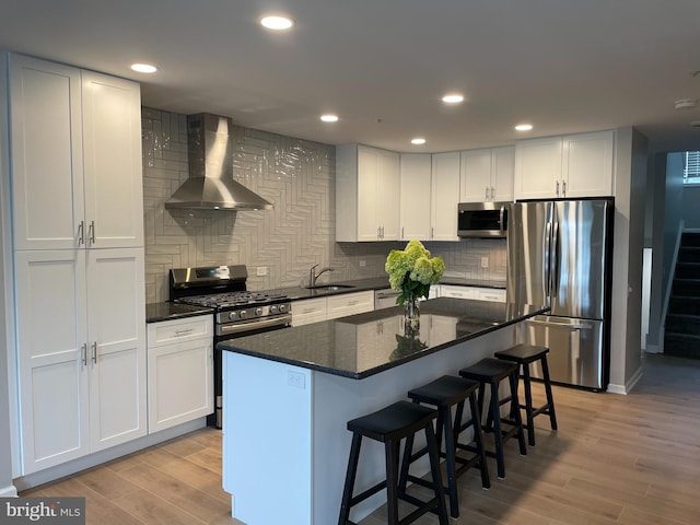 kitchen with wall chimney range hood, appliances with stainless steel finishes, white cabinetry, light wood-type flooring, and backsplash