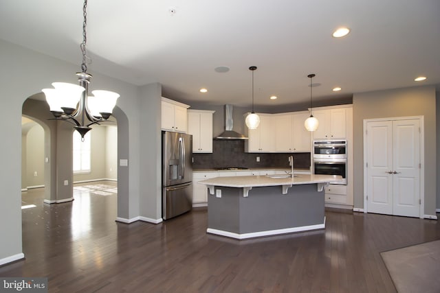 kitchen featuring appliances with stainless steel finishes, arched walkways, a sink, white cabinets, and wall chimney exhaust hood
