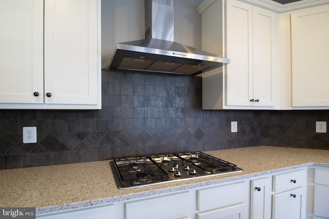 kitchen with wall chimney range hood, decorative backsplash, light stone counters, stainless steel gas stovetop, and white cabinetry