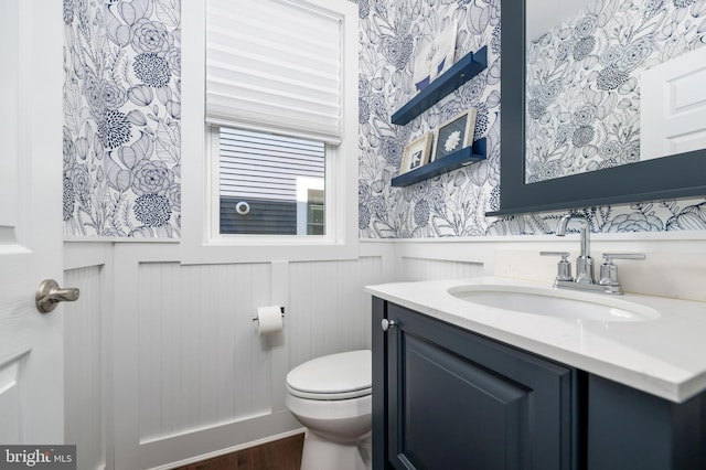 bathroom featuring wainscoting, vanity, toilet, wallpapered walls, and wood finished floors