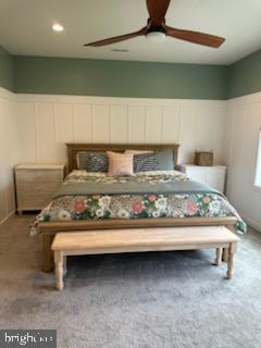 carpeted bedroom with a ceiling fan, wainscoting, and a decorative wall