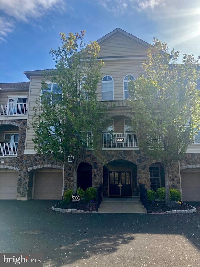 view of property featuring driveway, french doors, a balcony, an attached garage, and stone siding