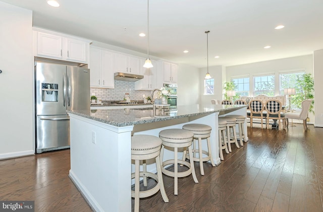 kitchen featuring decorative backsplash, dark wood-style floors, under cabinet range hood, stainless steel fridge with ice dispenser, and light stone counters