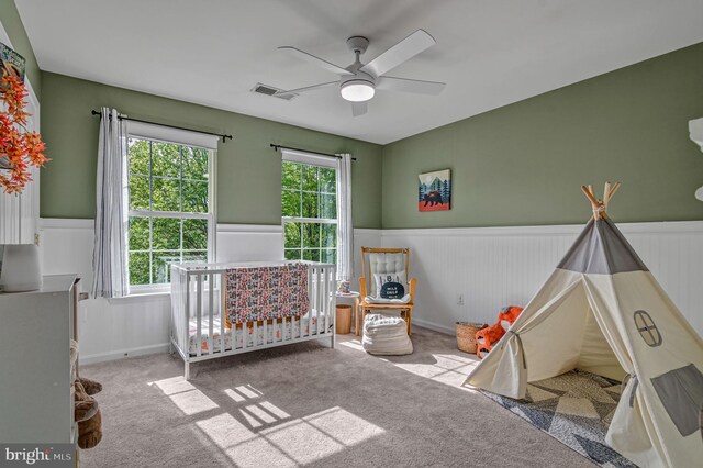 carpeted bedroom featuring wainscoting, multiple windows, a crib, and ceiling fan