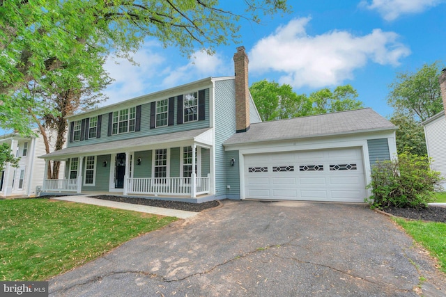 colonial home featuring aphalt driveway, a garage, a front yard, a chimney, and covered porch