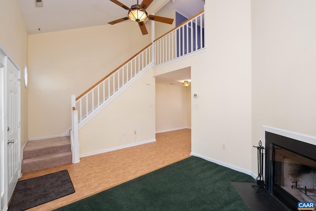 unfurnished living room featuring a ceiling fan, stairs, a fireplace with flush hearth, a high ceiling, and carpet flooring
