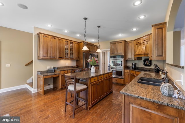 kitchen featuring appliances with stainless steel finishes, a sink, brown cabinetry, premium range hood, and a kitchen island