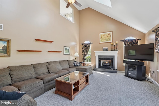 carpeted living room featuring a healthy amount of sunlight, high vaulted ceiling, a ceiling fan, and a warm lit fireplace